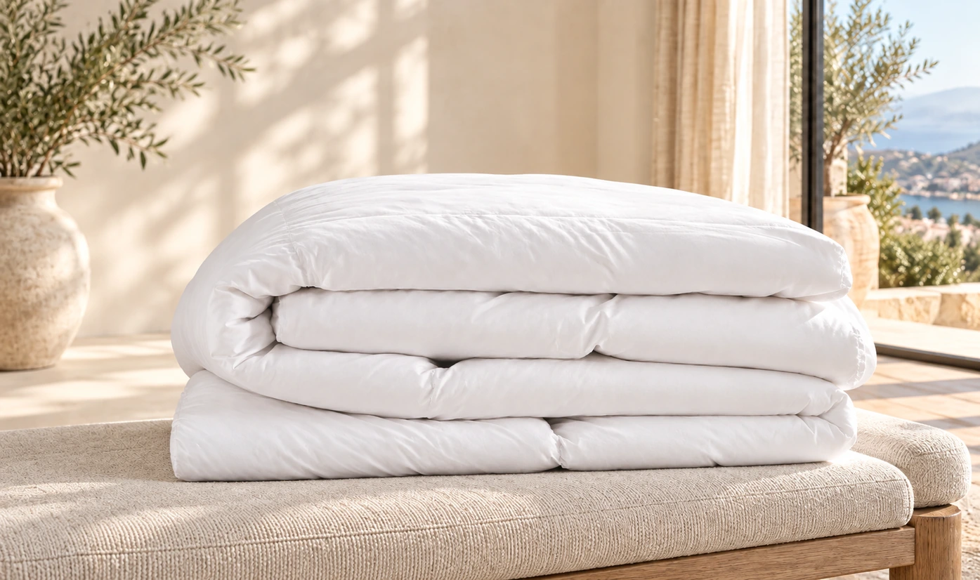 Folded white quilted comforter stacked on a beige textured blanket near a sunlit window